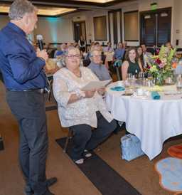 Chris Askin stands with a mic as Margaret Stewart and her family look on from their seats at their table.