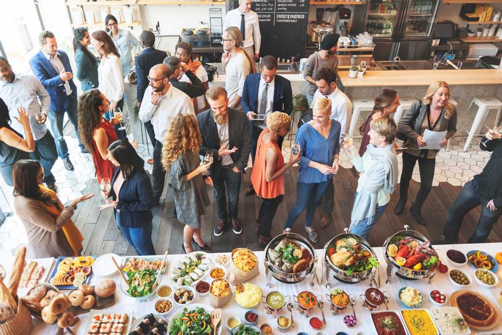 A group of about 30 adults gathered at an event, near a table full of appetizers and refreshments.