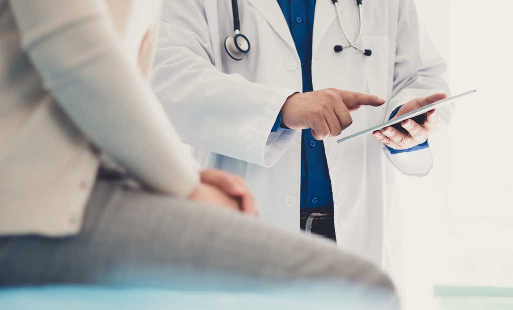 A male doctor visits with a seated woman patient, cropped so only below their shoulders is viewable.