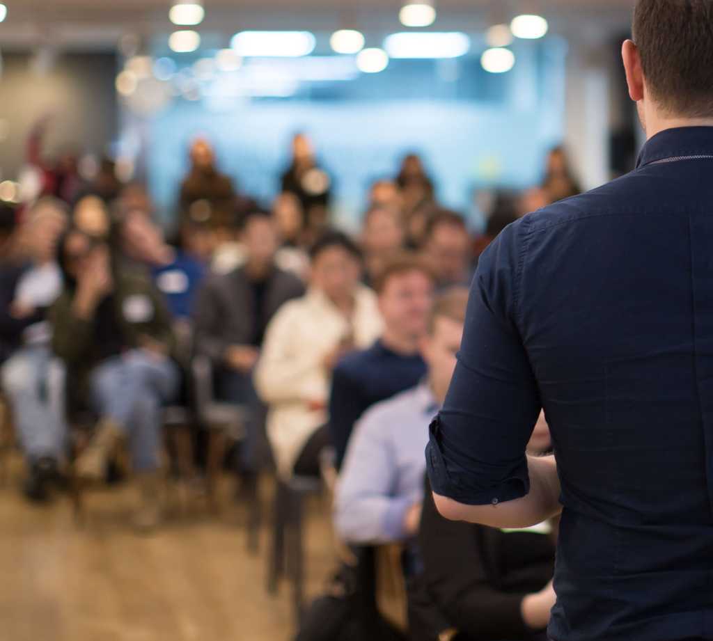 Over the shoulder view of a male presenter looking out into seated audience in a room.